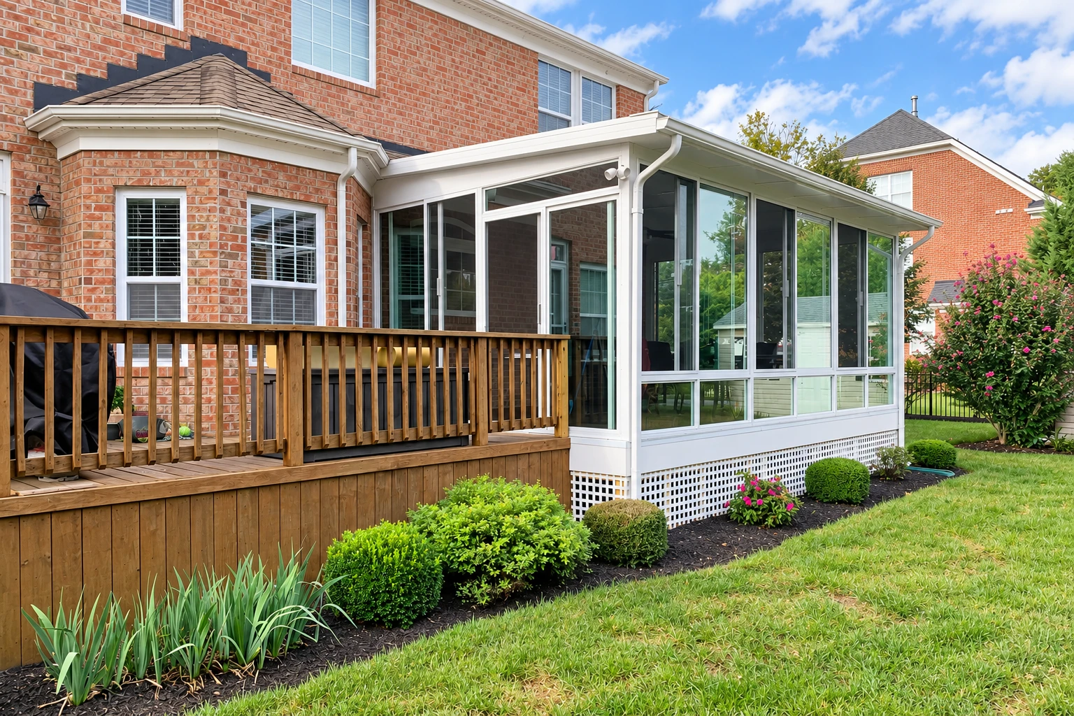 Glass sunroom addition beside a brick house, adjoining a raised wooden deck and landscaped lawn.