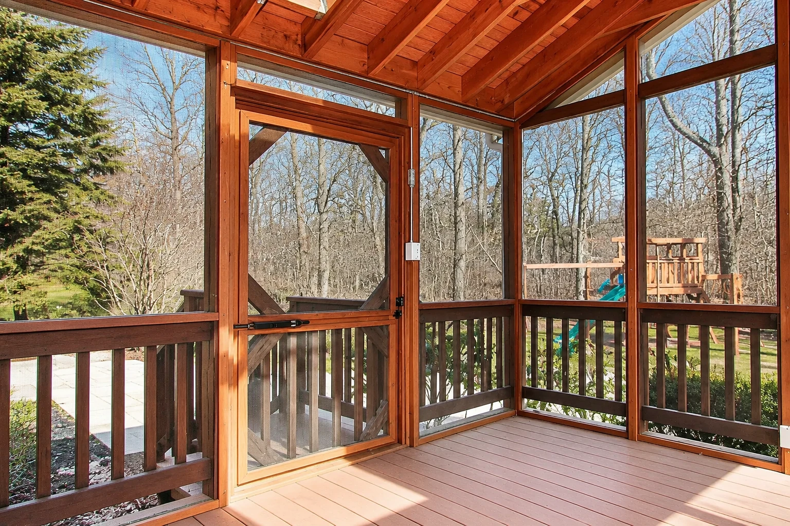 A wood-framed screen room has a mesh door and windows, with a timber floor and bare trees visible outside.