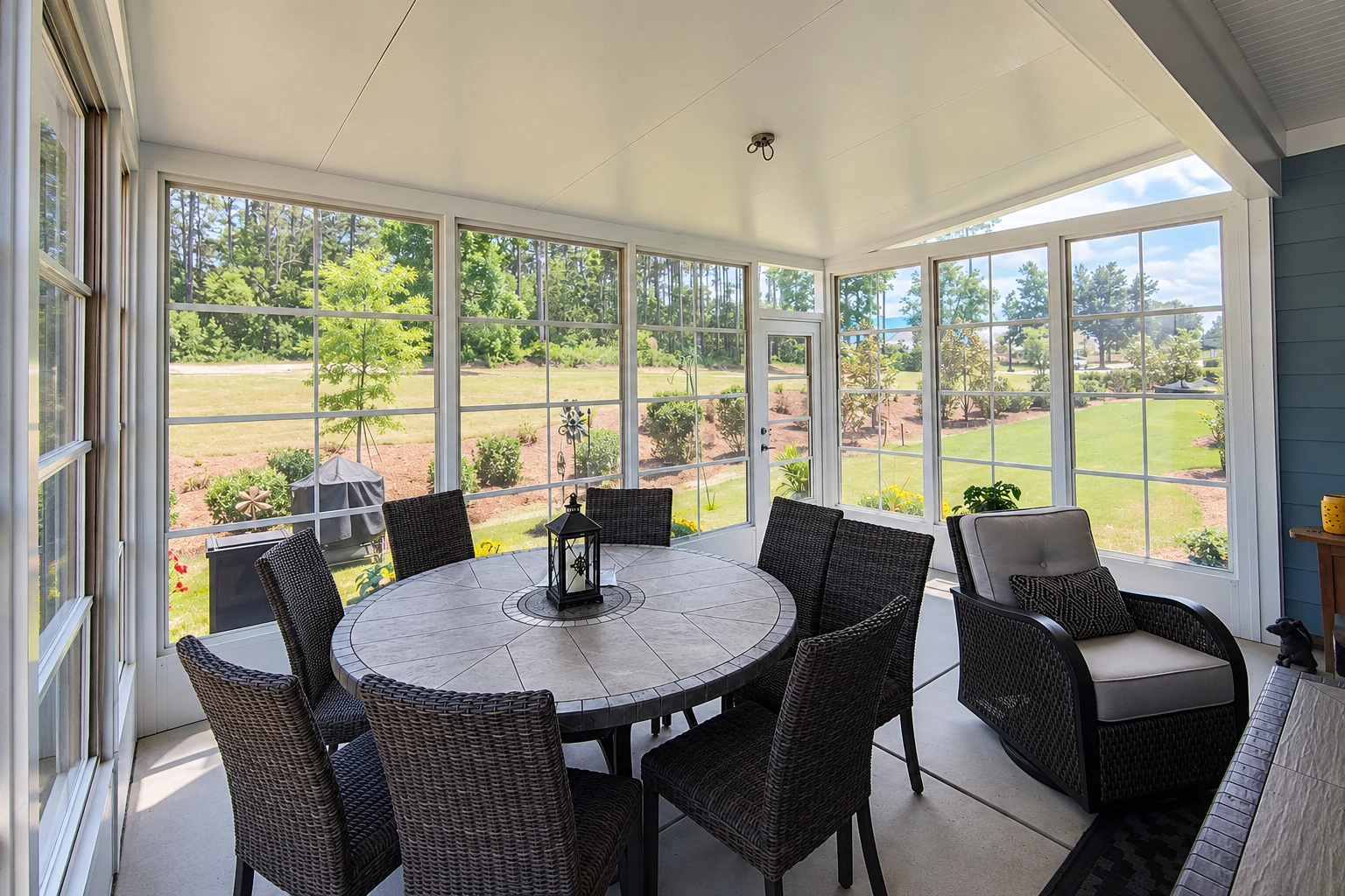 A bright sunroom addition with white-framed windows, a round stone-look table, and dark wicker chairs overlooking a lawn.
