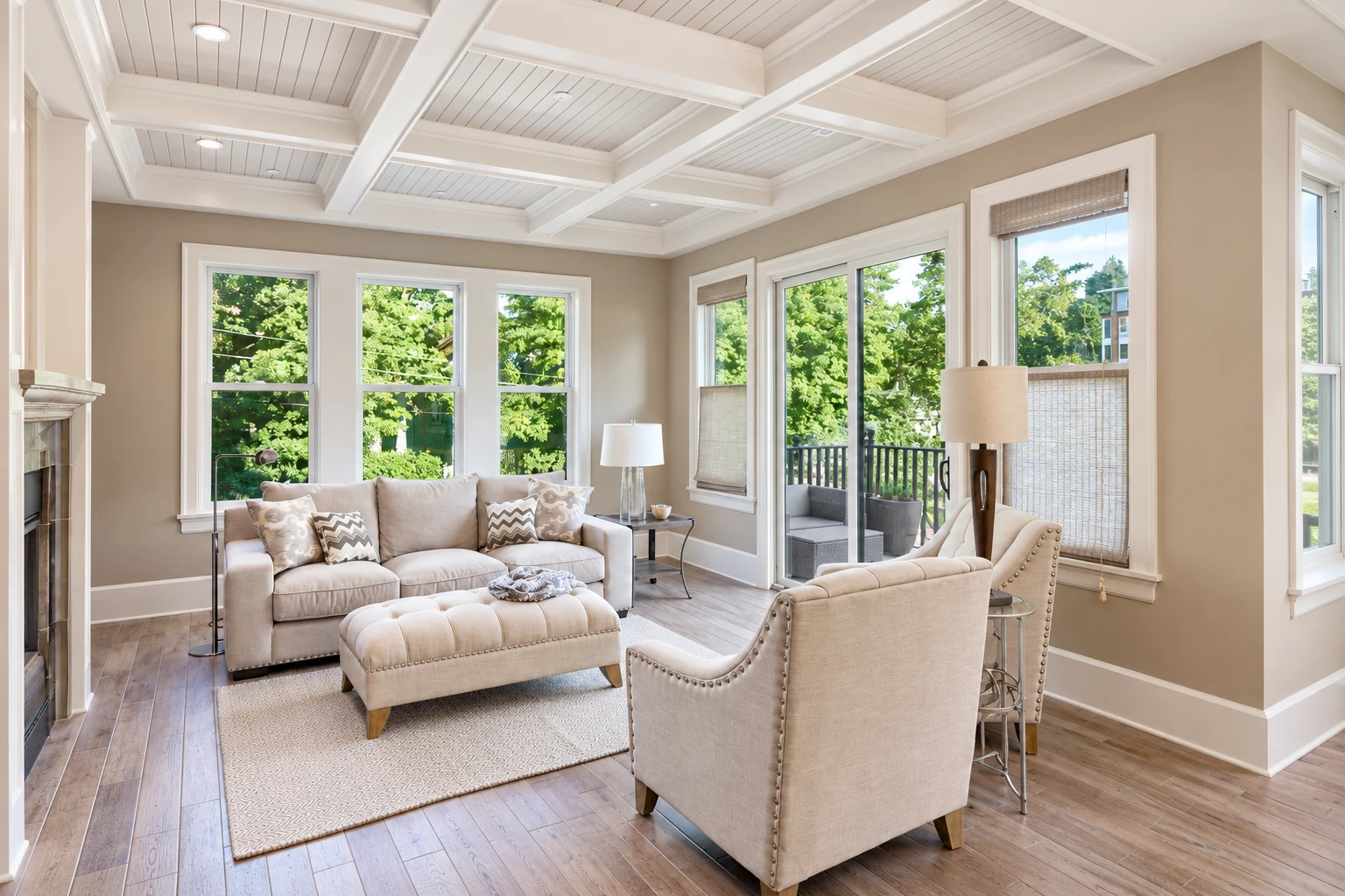 A bright sunroom with a white beamed ceiling, beige seating, and large windows overlooking leafy trees.