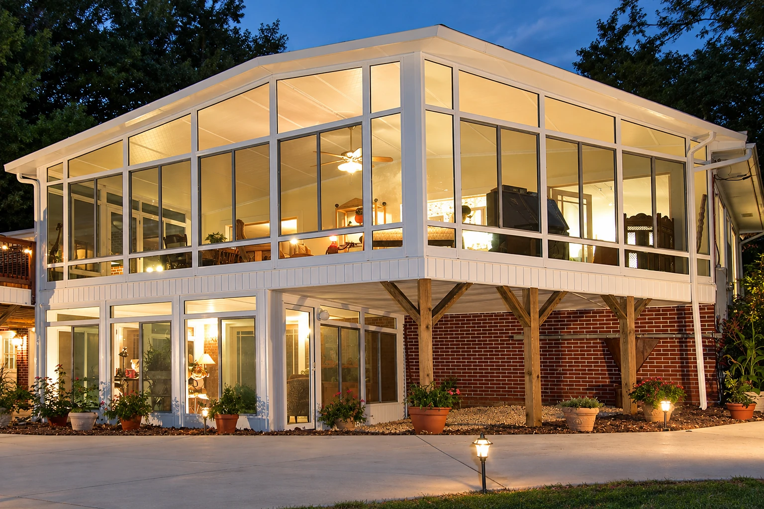 A two-story enclosed sunroom with white-framed glass walls glows warmly against the dark evening sky.