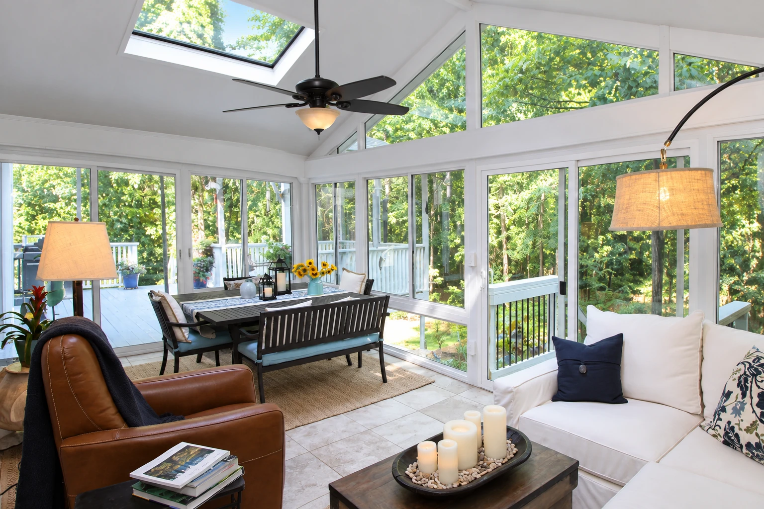 Bright three-season sunroom with expansive windows, a skylight, dining table, and white seating overlooking trees.